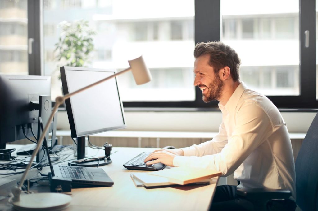 pexels photo 840996 A man smiling while working at an office desk with a computer and natural daylight streaming in through large windows.